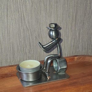 May include: A metal sculpture of a person sitting on a nut, holding a tea light candle holder. The sculpture is made from recycled metal parts and has a rustic, industrial look.