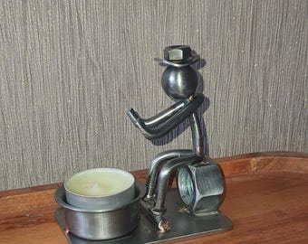 May include: A metal sculpture of a person sitting on a nut, holding a tea light candle holder. The sculpture is made of recycled metal parts and has a rustic, industrial look.