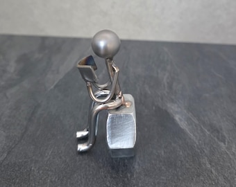 Puede incluir: Pequeña escultura metálica de una persona sentada leyendo. La figura es de metal color plata, con una cabeza esférica y una base rectangular. La persona sostiene un libro con las páginas abiertas.