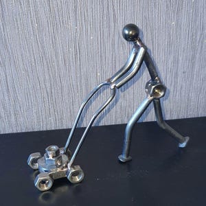 May include: A metal sculpture of a person pushing a lawnmower. The sculpture is made of nuts, bolts, and other metal pieces. The lawnmower has four wheels and a handle.