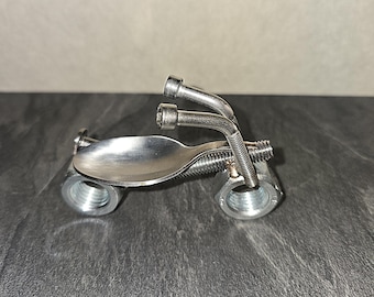 May include: A handcrafted metal sculpture of a motorcycle. The motorcycle is constructed from various metal objects, including a spoon for the seat, bolts, nuts, and springs. The handlebars are made from bent metal rods. The sculpture is silver in color.