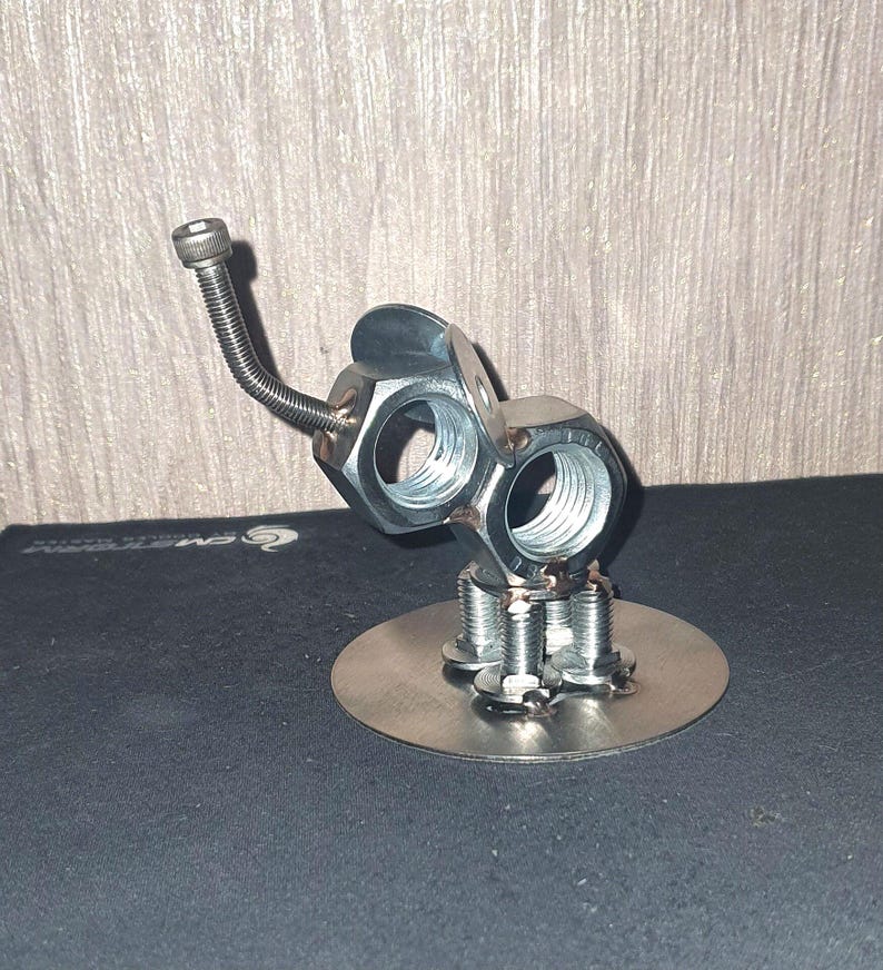 May include: A silver metal sculpture of an elephant made from nuts and bolts. The elephant has a long trunk and is standing on a round base.