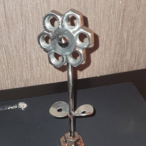 以下が含まれることがあります： ナットとボルトでできた銀色の金属製のフラワー彫刻。花は6枚の花びらと2枚の葉を持つ茎を持ちます。茎の基部は単一のナットです。