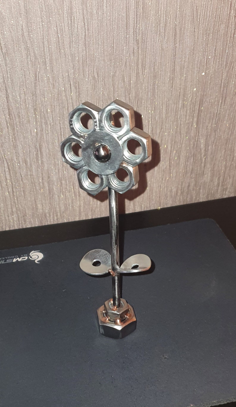 以下が含まれることがあります： ナットとボルトでできた銀色の金属製のフラワー彫刻。花は6枚の花びらと2枚の葉を持つ茎を持ちます。茎の基部は単一のナットです。