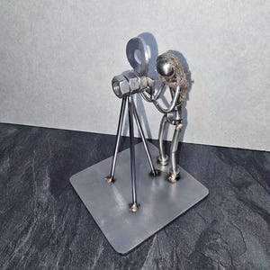 May include: A metal sculpture depicting a person filming with a vintage movie camera. The camera is made from nuts and bolts, with a circular film reel. The figure has a metal head and wire hair, standing on a square metal base.
