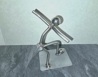 May include: A silver metal sculpture of a person lifting weights. The figure is made from various metal pieces, including a ball for the head, and a square base. The barbell is held overhead. The sculpture is on a dark gray surface.