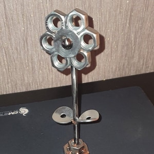 以下が含まれることがあります： ナットとボルトでできた金属製のフラワー彫刻。花は銀色の仕上げで、茎に葉が2枚ついています。彫刻のベースは大きなナットです。