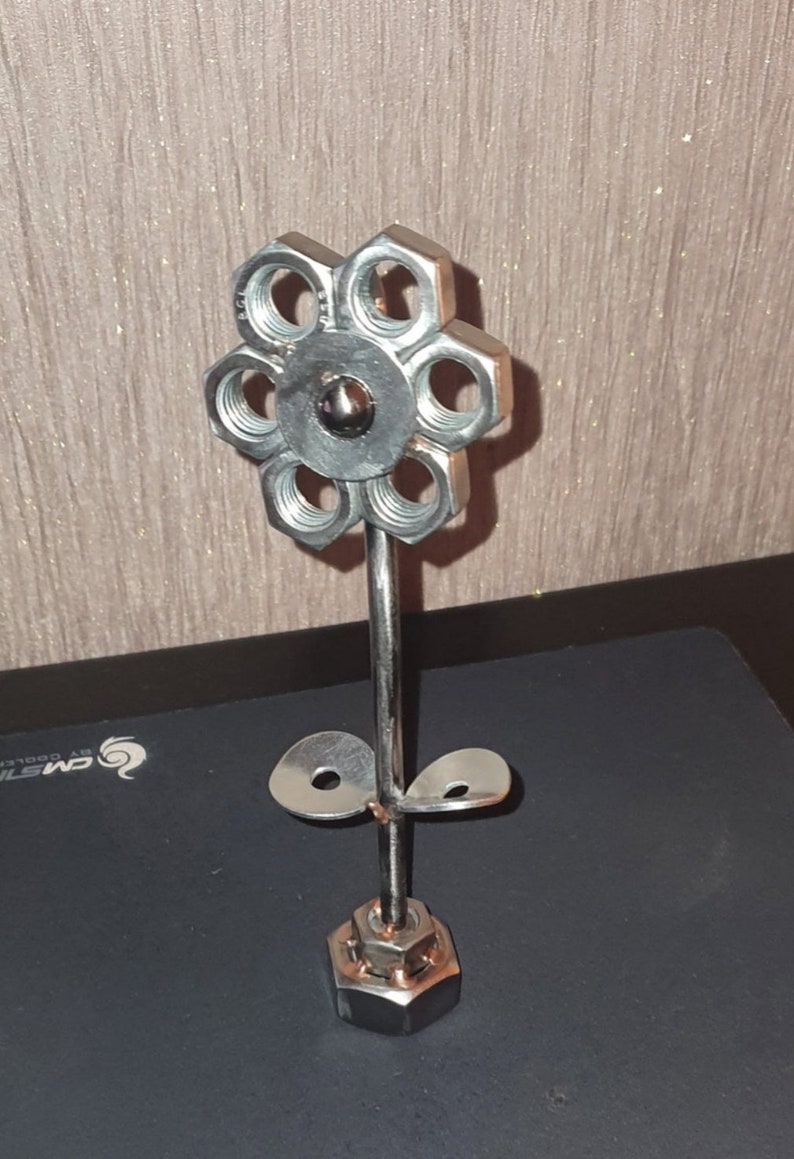 以下が含まれることがあります： ナットとボルトでできた金属製のフラワー彫刻。花は銀色の仕上げで、茎に葉が2枚ついています。彫刻のベースは大きなナットです。