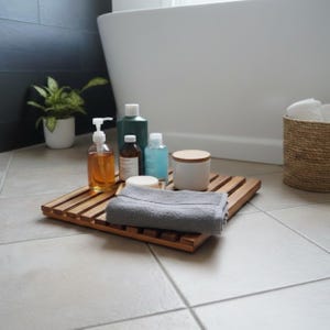 May include: A wooden bath tray holds various bath products, including a soap dispenser, bottles, and a gray towel. The tray is placed on a tiled floor, with a white bathtub in the background. A small potted plant and a woven basket are also visible.