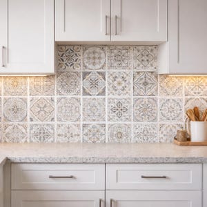 May include: Kitchen backsplash featuring decorative square tiles with intricate patterns in shades of gray, beige, and white. The tiles are installed above a countertop and below white cabinets. The kitchen design has a neutral color palette.