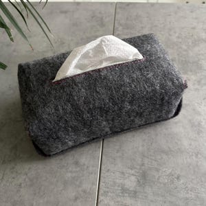 May include: A rectangular, dark grey felt tissue box cover with a white tissue protruding from the top opening. The opening is trimmed with a thin, dark red stitch. The tissue box cover has a simple, modern design.