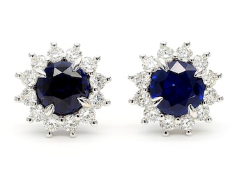 May include: A pair of stud earrings featuring a large, round, deep blue gemstone surrounded by a halo of smaller, clear gemstones. The earrings are set in a silver-colored metal, creating a sparkling, elegant design. The earrings are displayed against a white background.