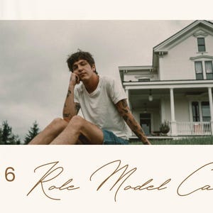 May include: A 2026 calendar with the text "Role Model Calendar" in a cursive font. The image shows a person sitting on grass in front of a house. The person is wearing a white t-shirt and denim shorts.