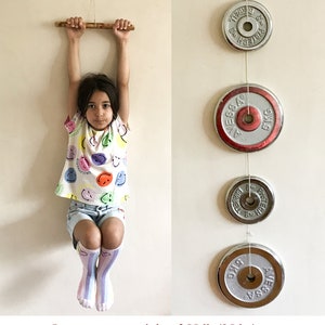 May include: A young person hangs from a wooden bar, with weight plates suspended nearby. The plates are silver with red accents and are labeled with weight measurements. The text in the image states the bar can carry 55 lb (25 kg).
