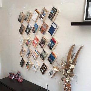 May include: A collection of books mounted on a white wall, arranged in a diamond pattern. The books have various covers, and some are held in place with small clips. A decorative vase with dried flowers and a dark brown cabinet are also visible.