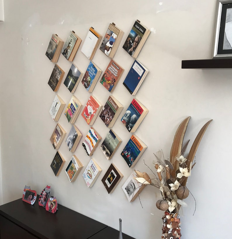 May include: A collection of books mounted on a white wall, arranged in a diamond pattern. The books have various covers, and some are held in place with small clips. A decorative vase with dried flowers and a dark brown cabinet are also visible.