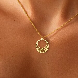 Puede incluir: Un collar dorado con un colgante circular con un dise&ntilde;o floral. El colgante cuelga de una delicada cadena. El collar se lleva sobre la piel, con una parte superior estampada en blanco y negro visible.