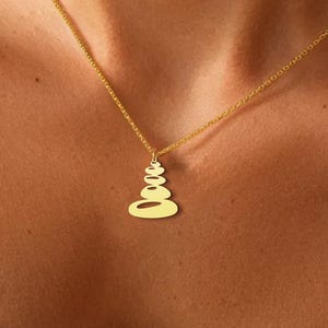 K&ouml;nnte beinhalten: Eine goldfarbene Halskette mit einem Anh&auml;nger in Form von gestapelten Steinen. Der Anh&auml;nger h&auml;ngt an einer feinen Kette. Die Halskette wird auf der Haut getragen, mit einem schwarz-wei&szlig; gemusterten Oberteil.