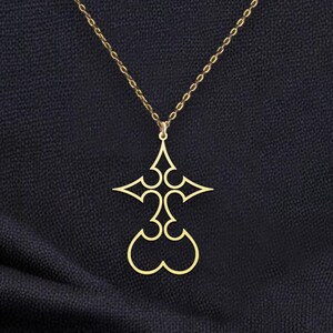 Puede incluir: Collar con colgante dorado con un dise&ntilde;o de cruz estilizada. El colgante cuelga de una delicada cadena sobre un fondo oscuro. La cruz tiene cortes sim&eacute;tricos y una base en forma de coraz&oacute;n.