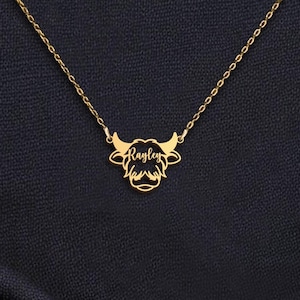 May include: A gold-colored necklace with a pendant shaped like a bull's head. The bull's head has horns and a detailed design. The name 'Raylen' is written in cursive across the center of the bull's head. The necklace is displayed on a dark blue fabric.