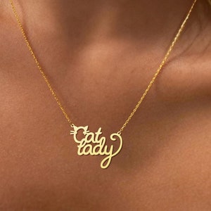 Peut inclure: Un collier en or avec un pendentif qui dit "Cat Lady" en &eacute;criture cursive. Le pendentif est en forme de t&ecirc;te de chat.