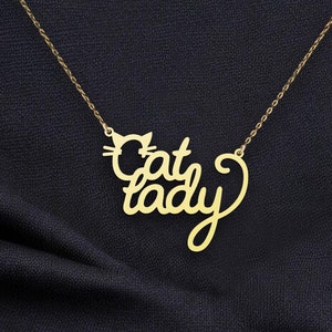 May include: A gold necklace with a pendant featuring the words 'Cat Lady' in a stylized script font. A small cat silhouette is incorporated into the design. The necklace is shown against a dark gray fabric background. The pendant is made of gold metal and has a delicate, feminine design. The chain is a fine, delicate gold chain.