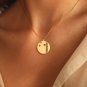 May include: A gold-colored pendant necklace with a circular pendant. The pendant features a design with three dots and a vertical stripe. The necklace is worn on a person's neck, and the chain is delicate.
