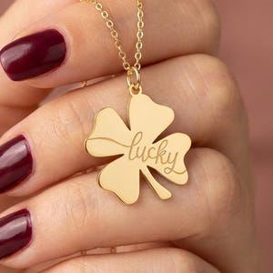 May include: A gold-colored necklace featuring a four-leaf clover pendant. The pendant has the word "lucky" engraved on it. The necklace is held up against a blurred background. The person holding the necklace has burgundy nail polish.