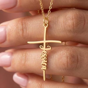 May include: A gold-coloured cross pendant necklace with the name "Jessica" inscribed vertically. The cross has a horizontal bar and a vertical bar, and the name is in a cursive font.