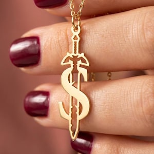 May include: A gold-colored pendant necklace featuring a sword design with a stylized letter "S" incorporated into the blade. The necklace is held by a person with burgundy nail polish. The chain is delicate and the pendant is intricately detailed.