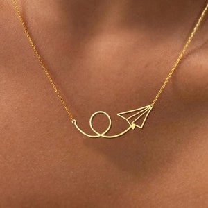 May include: A gold necklace with a delicate paper airplane pendant. The airplane is flying in a loop, symbolizing travel and freedom.