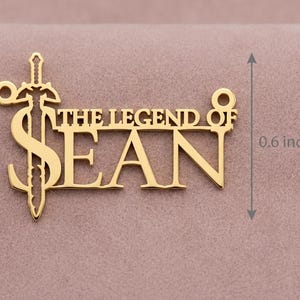 May include: A gold-colored pendant necklace with the text "THE LEGEND OF SEAN" and a sword design. The pendant is approximately 0.6 inches in height. The necklace is displayed on a pink surface.