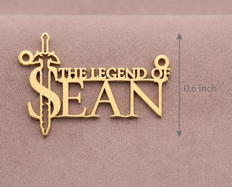 May include: A gold-colored pendant necklace with the text "THE LEGEND OF SEAN" and a sword design. The pendant is approximately 0.6 inches in height. The necklace is displayed on a pink surface.