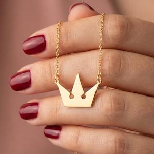 May include: A gold-colored crown pendant necklace. The crown is a simple, stylized design with three points. The necklace is held by a person with red nail polish. The chain is delicate and the pendant is centered.