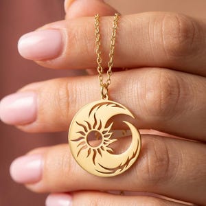 Puede incluir: Un collar con colgante dorado con un dise&ntilde;o de sol y luna creciente. El sol est&aacute; en el centro, con rayos que se extienden. La luna se curva alrededor del sol. El collar es sostenido por una persona.