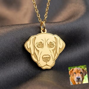 May include: A gold-toned metal necklace featuring a pendant shaped like a dog's head with engraved facial features. The pendant is suspended from a delicate gold-toned chain. A small square photograph of a brown dog is positioned in the lower right corner.