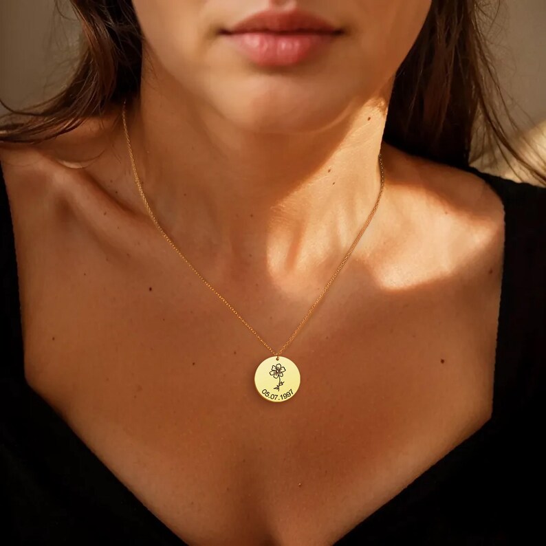 May include: A gold necklace with a circular pendant. The pendant has a floral design and the date 05.07.1997. The necklace is worn, hanging against a black top.