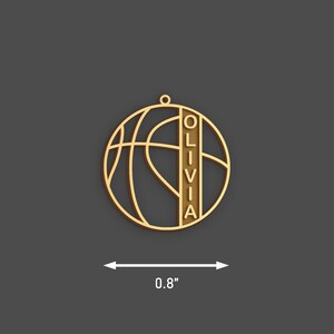 May include: A gold-colored basketball pendant with the name "OLIVIA" vertically displayed. The pendant is approximately 0.8 inches in diameter and features a basketball design. The pendant has a small loop at the top for hanging.