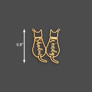 May include: Gold-colored cat-shaped pendant with the names "Shadow" and "Wendy" inscribed. The pendant is approximately 0.8 inches in height. The design features two cat silhouettes facing away, with a small loop at the top for hanging.
