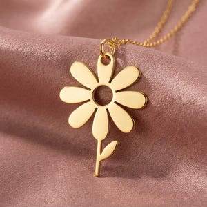 May include: A gold daisy pendant necklace. The pendant has a daisy flower design with a circular centre and petal cutouts. The necklace is displayed on a pink satin fabric.