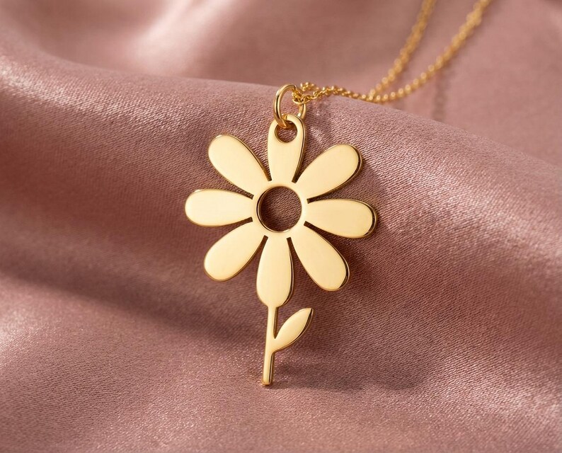 May include: A gold daisy pendant necklace. The pendant has a daisy flower design with a circular centre and petal cutouts. The necklace is displayed on a pink satin fabric.