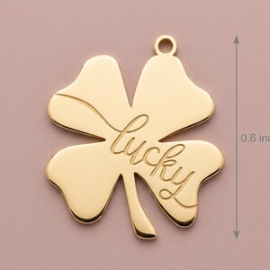 May include: A gold-colored four-leaf clover charm with the word "lucky" inscribed. The charm is approximately 0.6 inches in length and has a small loop at the top for attachment. The clover is set against a soft pink background.