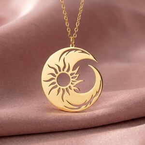 Puede incluir: Un collar con colgante dorado con un dise&ntilde;o de sol y luna creciente. El sol est&aacute; en el centro, con rayos que se extienden hacia afuera. La luna se curva alrededor del sol. El collar se muestra sobre una tela rosa.