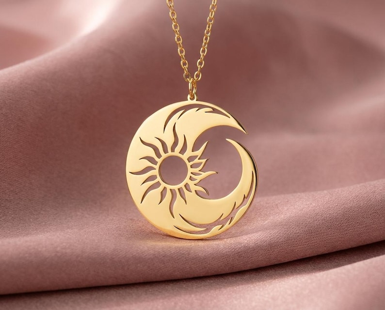 Puede incluir: Un collar con colgante dorado con un dise&ntilde;o de sol y luna creciente. El sol est&aacute; en el centro, con rayos que se extienden hacia afuera. La luna se curva alrededor del sol. El collar se muestra sobre una tela rosa.