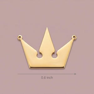 May include: A gold-colored crown pendant, approximately 0.6 inches in width, with a classic royal design. The crown features five points and two small holes at the top for attachment. The background is a muted mauve color.