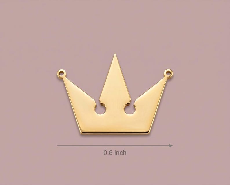 May include: A gold-colored crown pendant, approximately 0.6 inches in width, with a classic royal design. The crown features five points and two small holes at the top for attachment. The background is a muted mauve color.