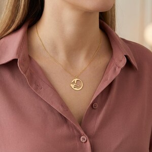 May include: A gold-colored pendant necklace featuring a circular design with a stylized figure. The necklace is worn on a person wearing a rose-colored button-down shirt. The pendant hangs from a delicate chain.