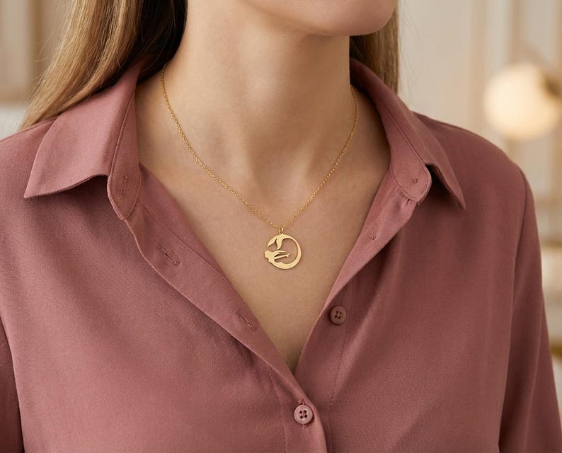 May include: A gold-colored pendant necklace featuring a circular design with a stylized figure. The necklace is worn on a person wearing a rose-colored button-down shirt. The pendant hangs from a delicate chain.
