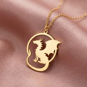 May include: A gold-colored pendant necklace featuring a dragon design. The dragon is inside a circular frame. The necklace is displayed on a pink satin fabric.