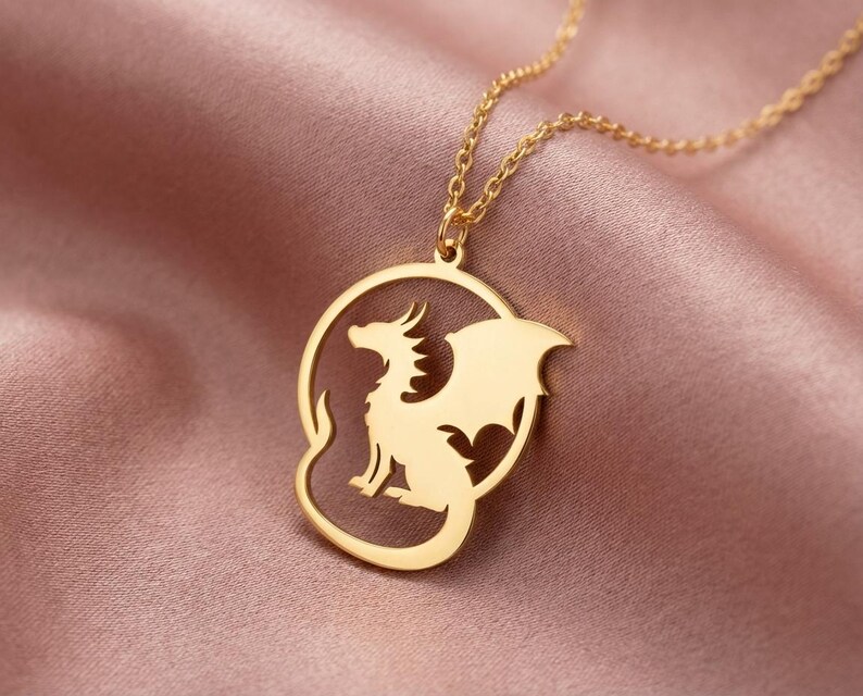 May include: A gold-colored pendant necklace featuring a dragon design. The dragon is inside a circular frame. The necklace is displayed on a pink satin fabric.